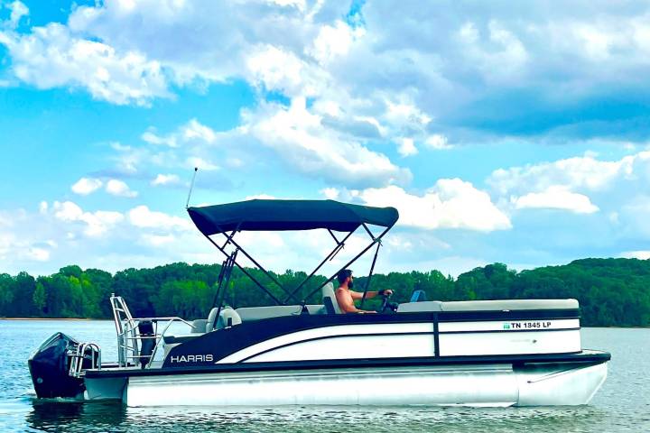 Man steering pontoon boat on lake with cloudy sky.