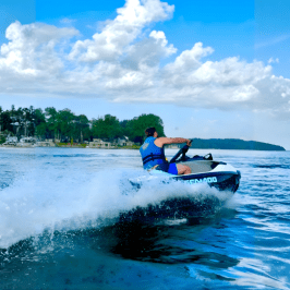 Person jet skiing on blue ocean near a tree-lined coastline under a cloudy sky.