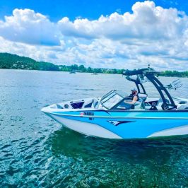 Person steering a blue and white speedboat on a lake, surrounded by trees and clouds.